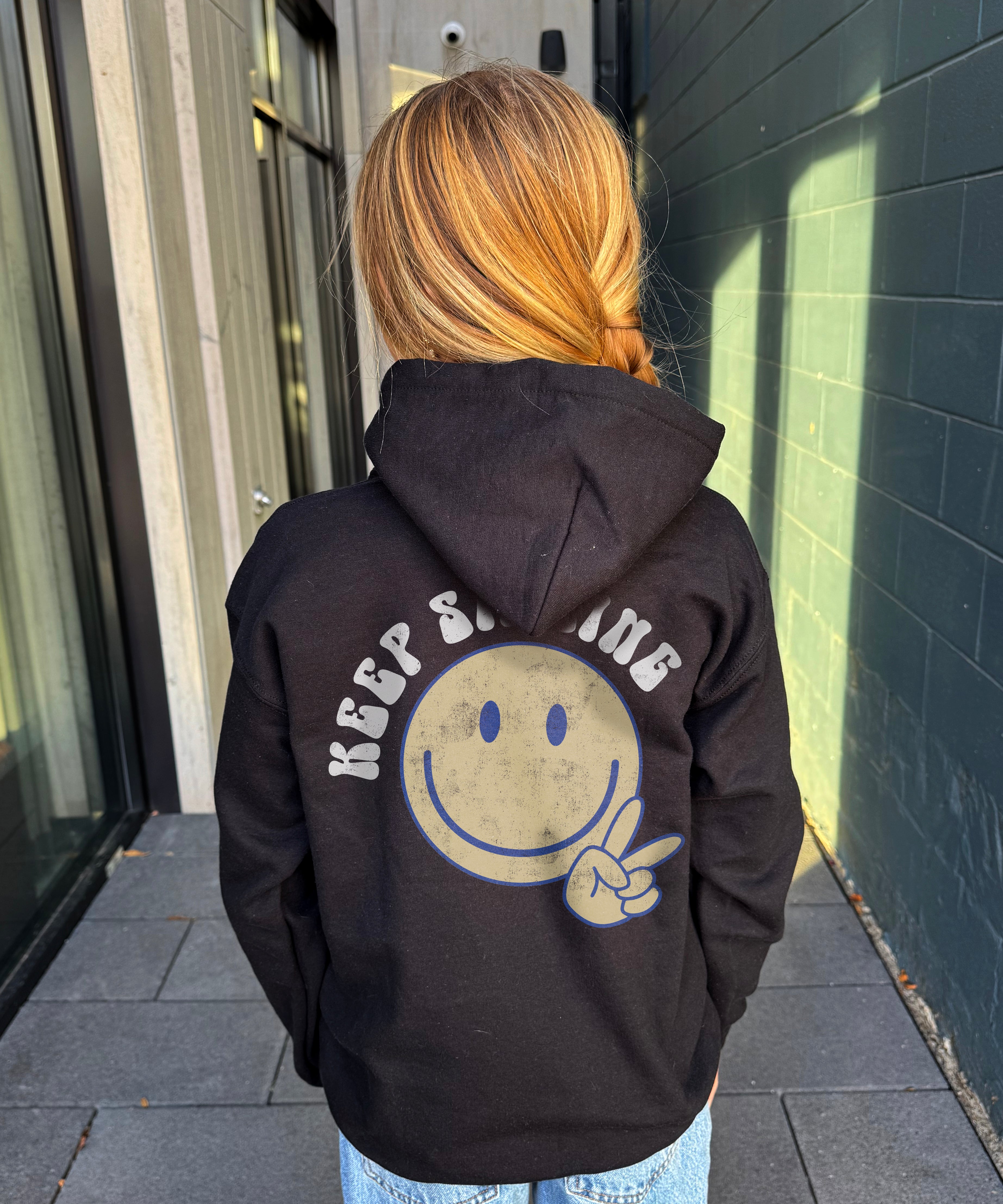 Girl in black hoodie that says " Keep smiling" in white text on the back with  smiley and peace sign