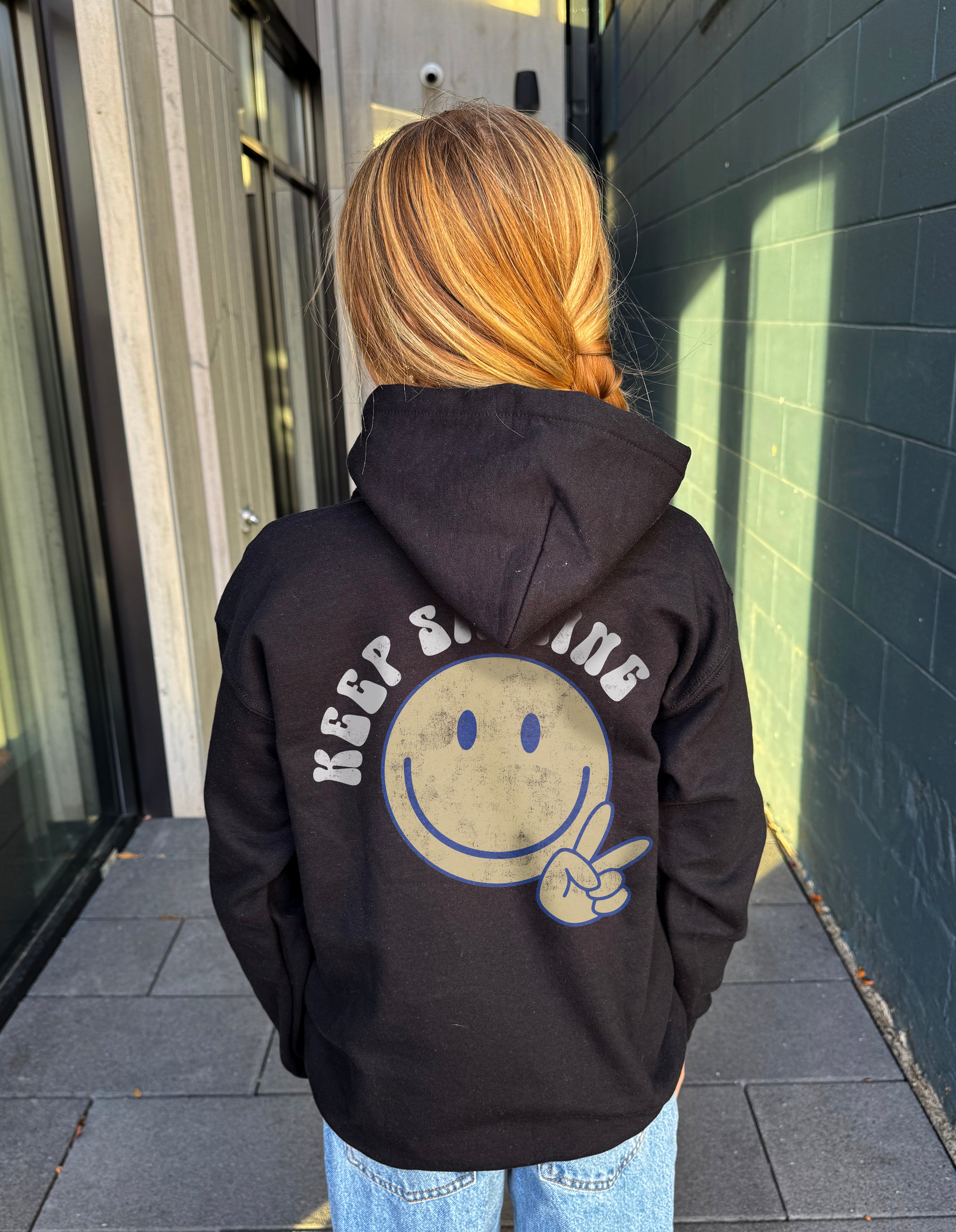 Girl in black hoodie that says " Keep smiling" in white text on the back with  smiley and peace sign