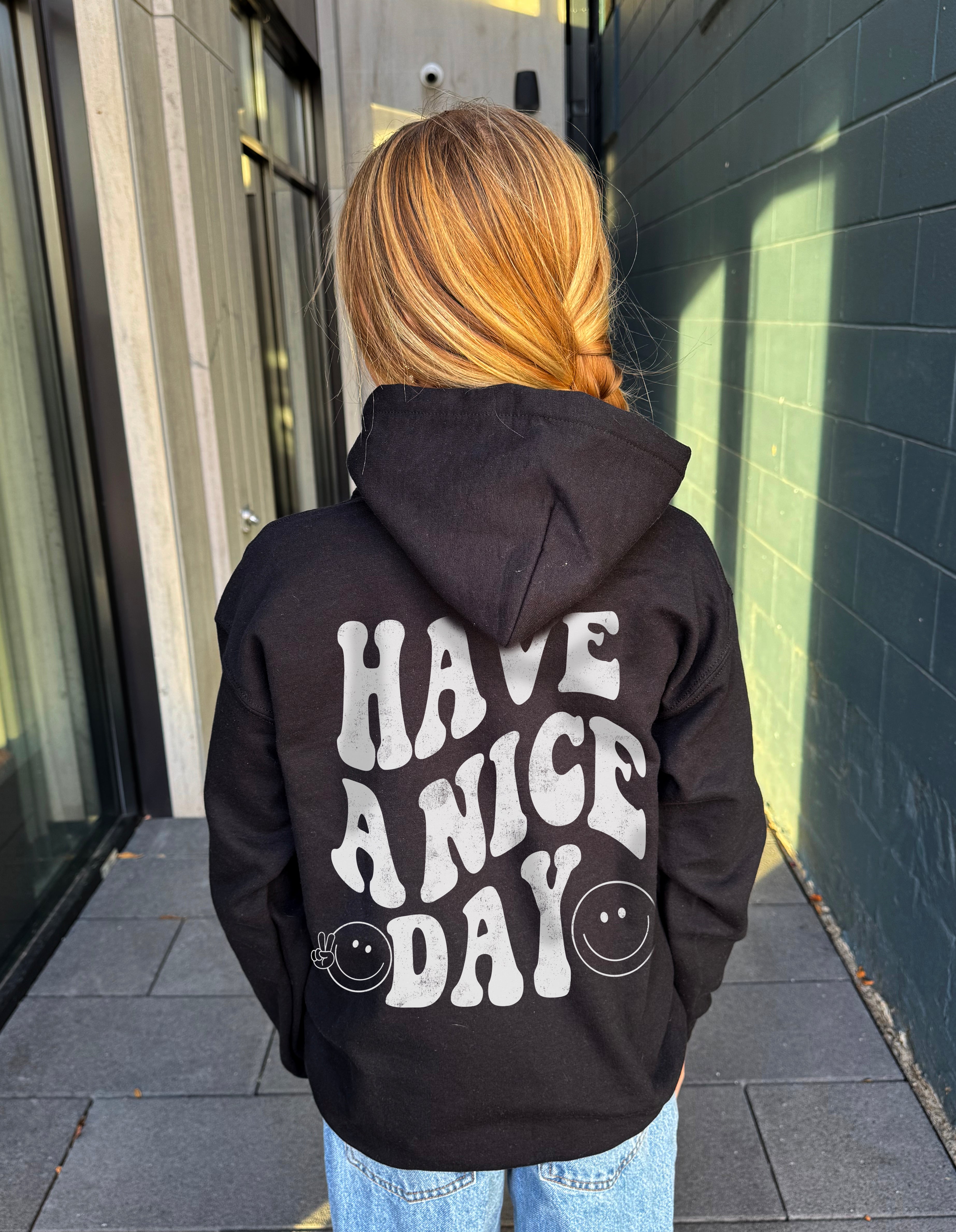 Girl in black hoodie that says "have a nice day" in white text with Smiley face from behind