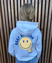 Girl in white hoodie that says " Keep smiling" in blue text on the back with  smiley and peace sign