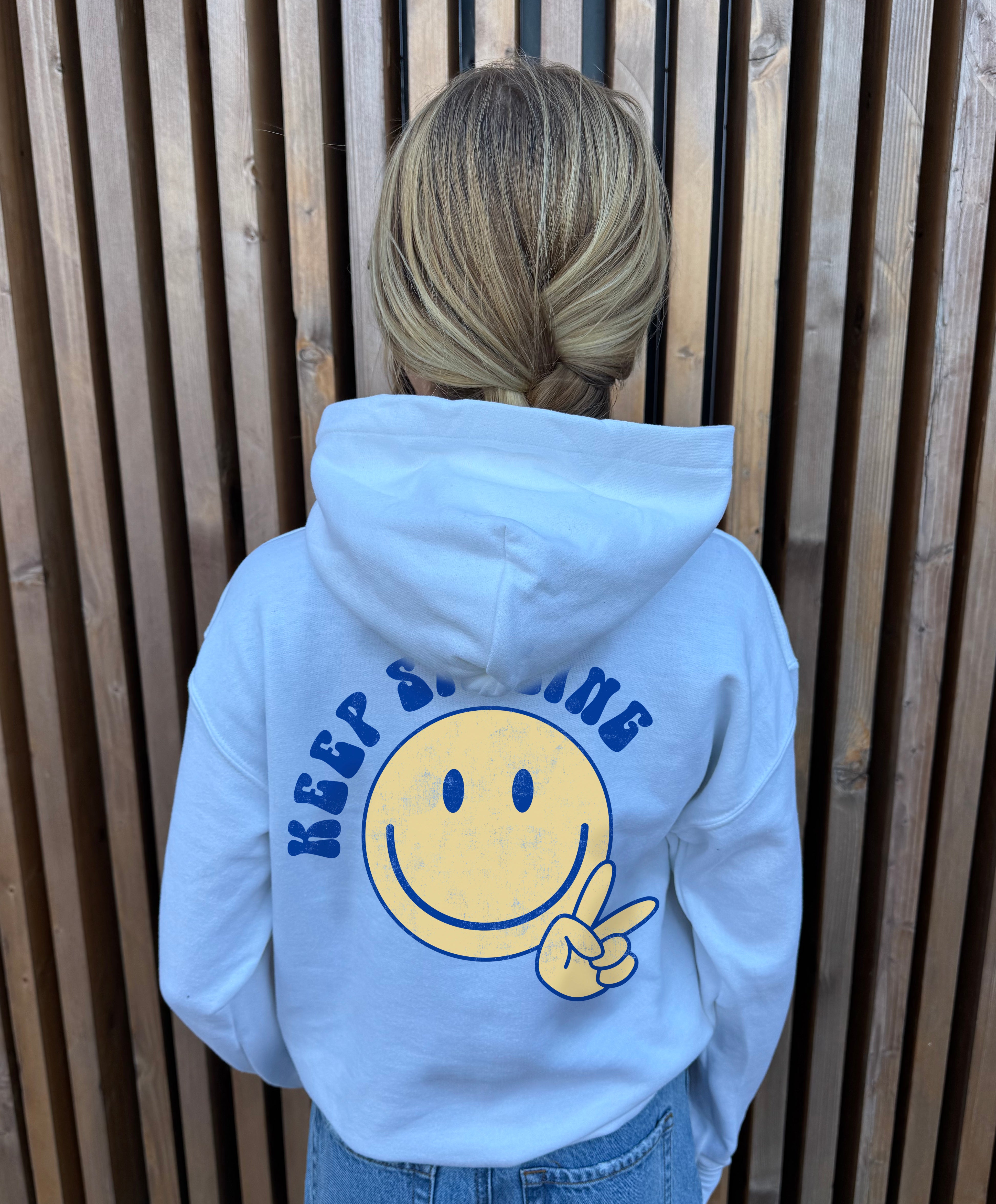 Girl in white hoodie that says " Keep smiling" in blue text on the back with  smiley and peace sign