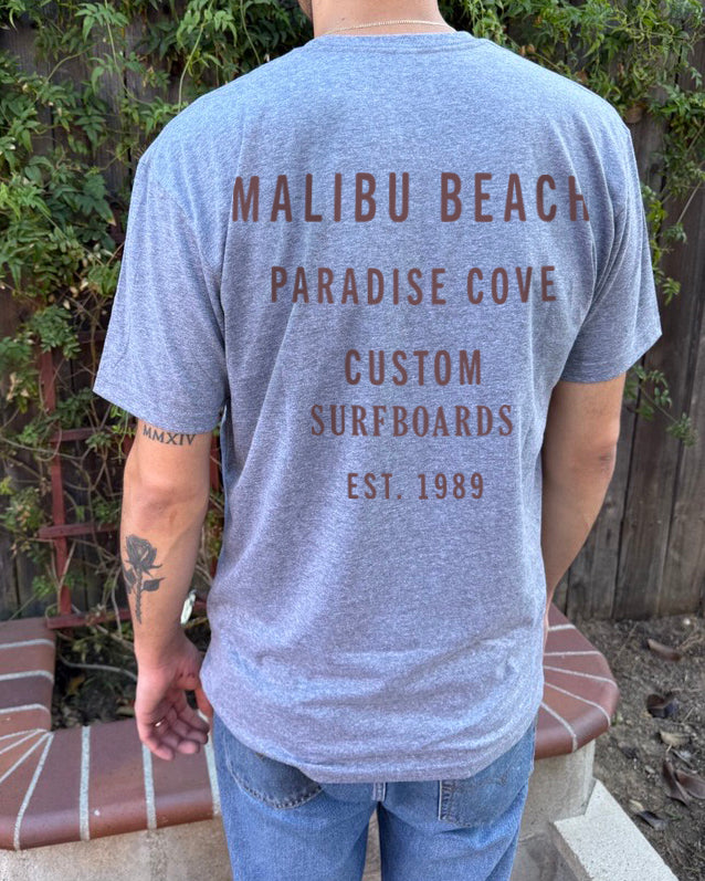 Boys t-shirt from the back in grey with rust red text that says "Malibu Beach Paradise Cove Custom Surfboards" 