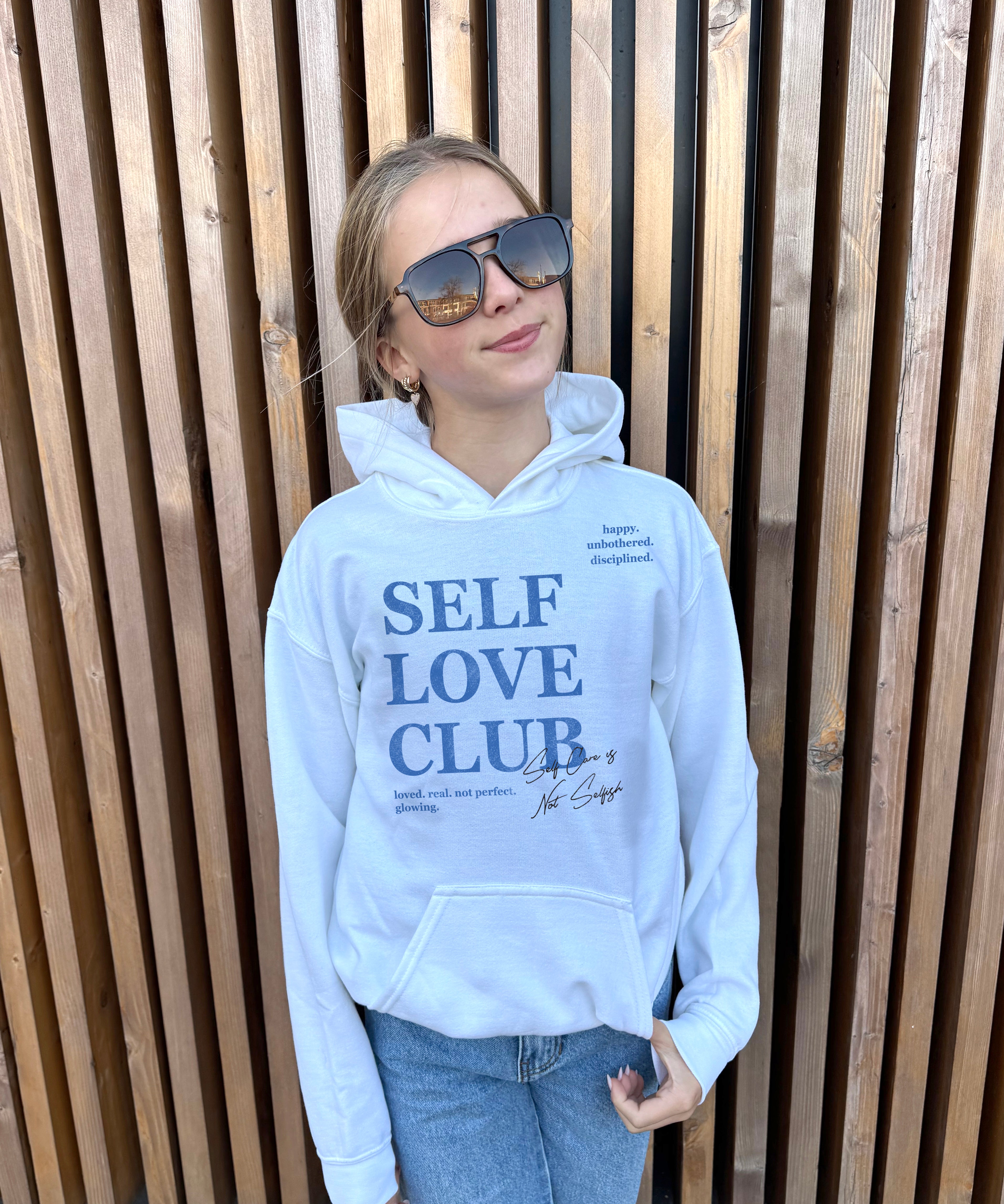 Girl in white hoodie that says "Self Love Club" in blue text 