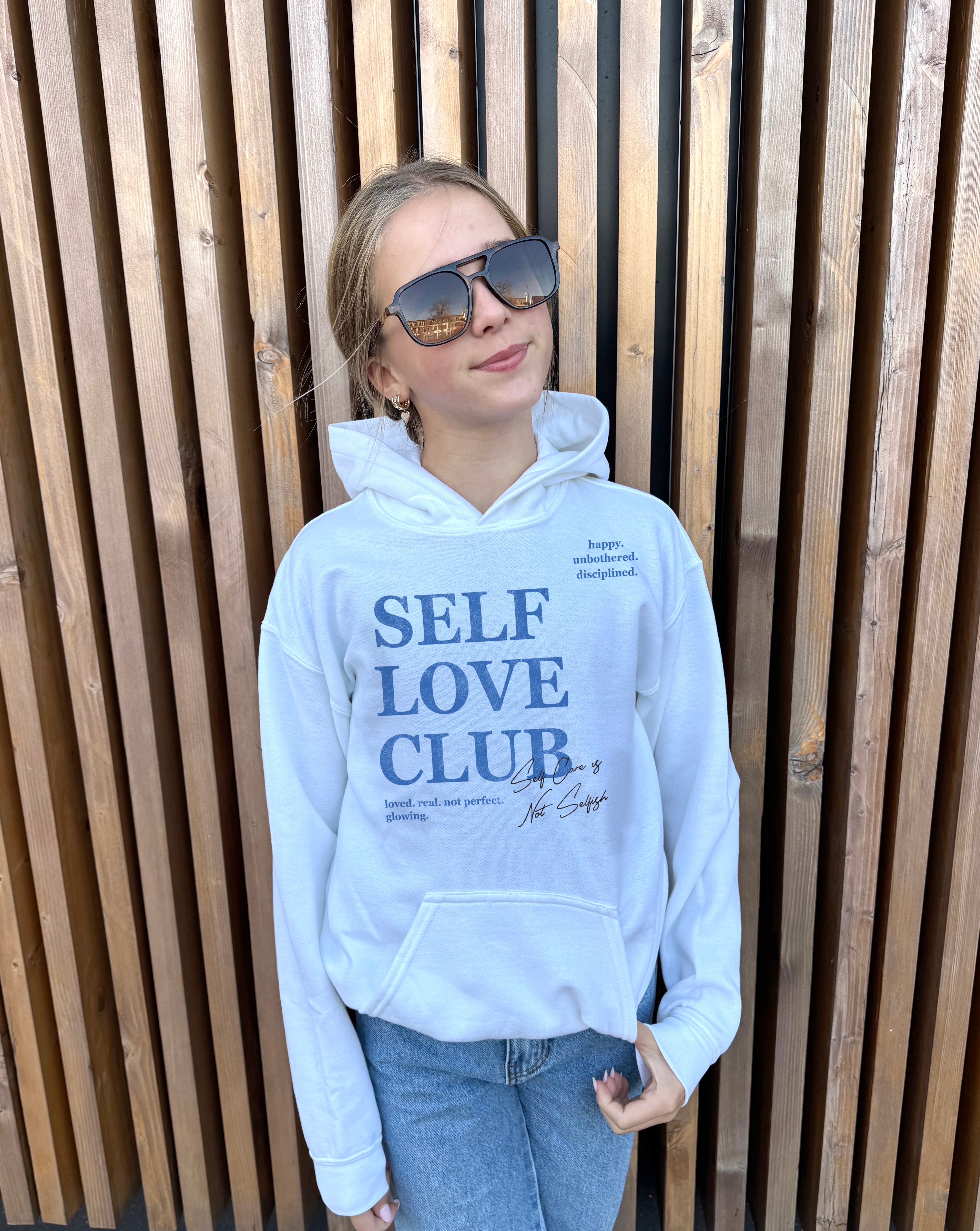 Girl in white hoodie that says "Self Love Club" in blue text 